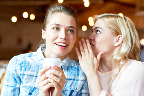 Attractive blond-haired woman sharing secret with friend while sitting at cozy small coffeehouse and enjoying cappuccino, blurred background