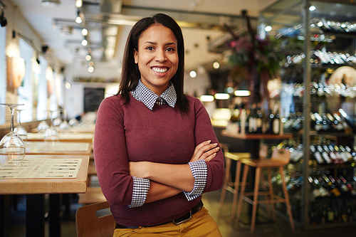 Young cross-armed female sitting in cafe with row of tables and bar counter behind