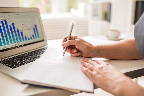 Close-up shot of busy manager taking necessary notes while analyzing statistics with help of modern laptop, blurred background