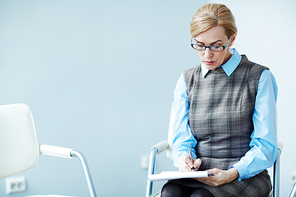 Portrait of mature woman wearing glasses sitting alone waiting for group therapy session to start and making notes