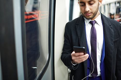 Young Middle-Eastern businessman standing by window in subway train using smartphone to listen to music and text messaging