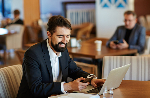 Waist-up portrait of smiling middle-aged businessman texting with his friend on modern smartphone while sitting in cafe and working on laptop