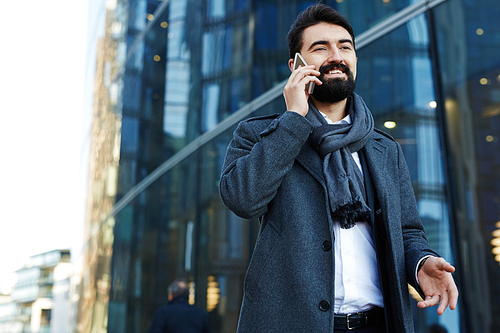 Low angle view of cheerful middle-aged businessman answering to phone call while standing at modern glass office building, waist-up portrait