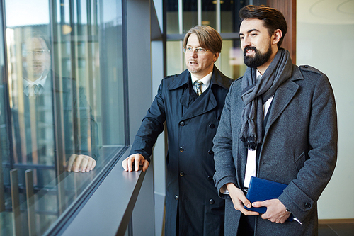 Confident middle-aged businessmen wearing coats standing in office lobby and looking out panoramic window, portrait shot