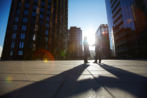 Low angle view of two unrecognizable businessmen shaking hands firmly while standing outdoors, last beams of setting sun illuminating them, lens flare