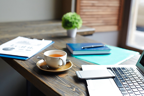 Closeup background shot of different business objects on table in cafe: laptop, smartphone, documents and folders next to cup of tea left by working businessman