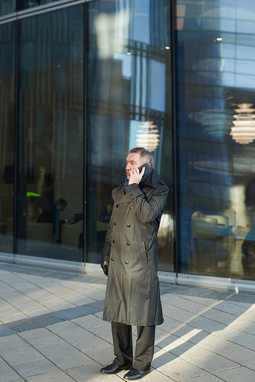 Full-length portrait of mature entrepreneur having phone conversation while standing at modern glass office building