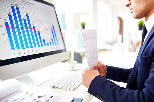 Modern financier with papers sitting in front of computer monitor with chart and graph of sales rate