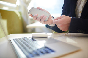 Unrecognizable businessman connecting his low battery smartphone to power bank while sitting at wooden cafe table with modern laptop, close-up shot