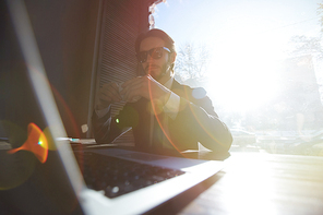 Portrait of cool man in business suit working with laptop in sunlit cafe, wearing big sunglasses and drinking coffee