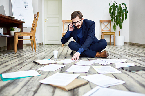 Portrait of middle aged bearded man speaking by phone and sorting documents sitting on floor in office