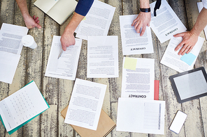 Top view of assorted documents and contracts laid out on wooden office floor with hands of two unrecognizable  people working