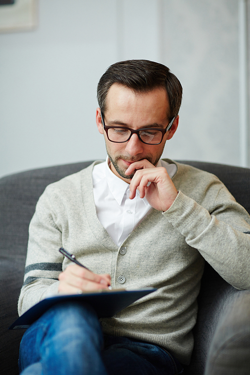 Pensive author or specialist sitting in armchair and making notes