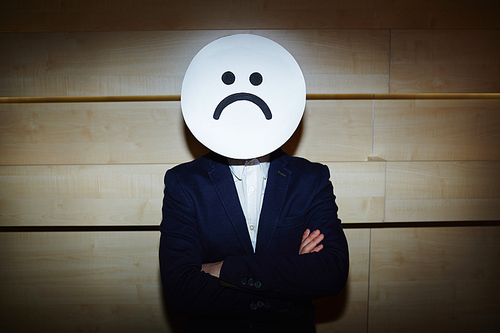 Upset businessman in white shirt and suit covered his face with sad mask while standing with arms crossed against wooden wall