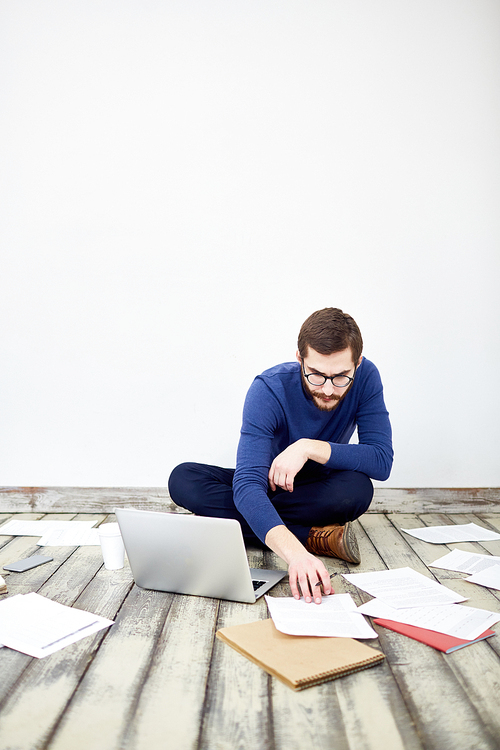 Portrait of bearded modern businessman sitting cross legged on floor in office,  working with laptop and sorting documentation laid out around