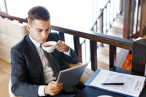 Young entrepreneur with touchpad drinking tea and looking through business news