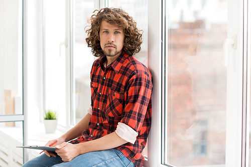 Young office worker in casual shirt and jeans holding pencil and clipboard with paper, leaning against window and  during work