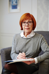 Portrait of mature psychologist in eyeglasses sitting on comfortable armchair with legs crossed and filling in medical card of patient