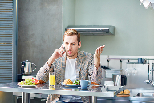 Confident young entrepreneur talking to his business partner on smartphone while having delicious breakfast at modern kitchen, waist-up portrait