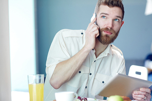 Portrait of stylish handsome man in casual shirt speaking by smartphone and using digital tablet while having lunch in cafe