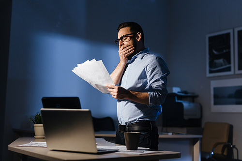 Portrait of tired bearded businessman wearing glassed yawning while working with documentation in dark office late at night, standing with heap of papers by desk