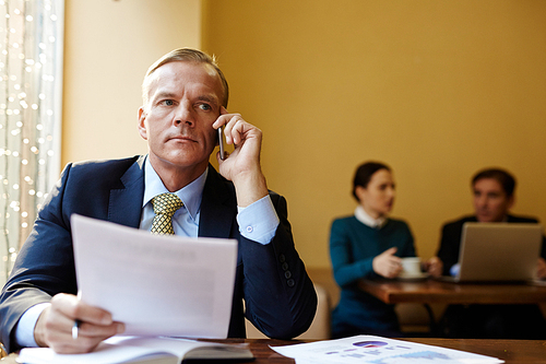 Serious employer speaking on mobile phone with two managers on background