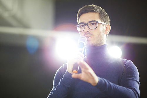 Head and shoulders portrait of confident young designer in eyeglasses holding microphone in hand while making presentation for his colleagues