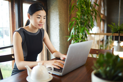 Asian businesswoman typing on laptop while sitting by table in modern office or cafe