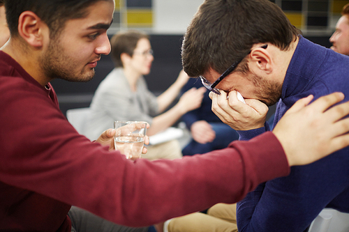 Careful friend supporting young man in difficult period of life and offering him glass of water