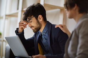 Side view portrait of exhaustd young middle eastern businessman pinching nosebridge looking at laptop screen sitting  with  female colleague helping him while working to meet important deadline together