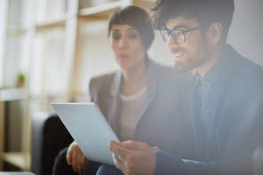 Young handsome middle eastern businessman wearing creative haircut and glasses smiling while looking at laptop screen and discussing project with colleague sitting in background behind him