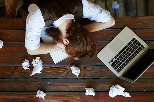 Tired designer sleeping on wooden table with crumpled papers around