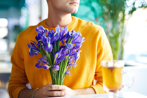 Portrait of unrecognizable young man holding bouquet of beautiful irises while waiting for date in cafe