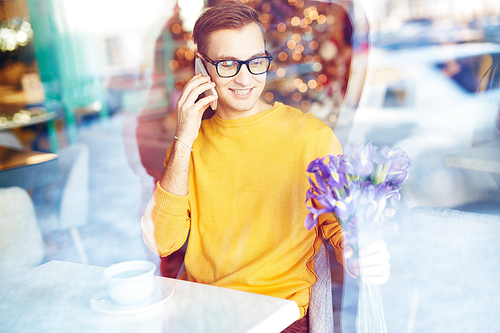 Portrait of smiling young man speaking by phone in cafe while waiting for date with bouquet  of irises