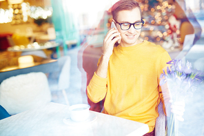 Portrait of smiling young man speaking by phone in cafe while waiting for date with bouquet  of flowers