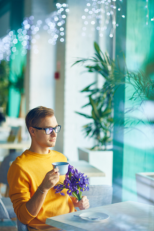 Portrait of trendy romantic man waiting for date in cafe with bouquet of flowers, drinking coffee