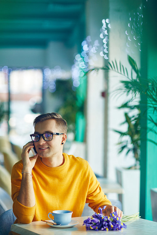 Portrait of smiling young man speaking by phone while waiting for romantic date in cafe with bouquet of flowers