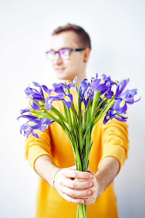 Portrait of smiling young man holding bouquet of flowers standing against white wall, focus on beautiful irises