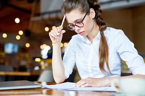 Concentrated young financial manager in eyeglasses preparing annual accounts while sitting at cafe table, blurred background