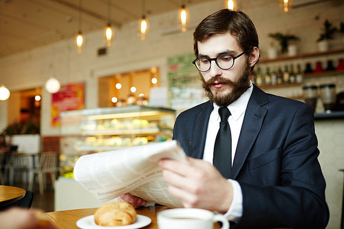 Well-dressed businessman in eyeglasses reading latest news by breakfast in cafe