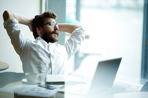 Relaxing businessman sitting in front of laptop and looking through office window