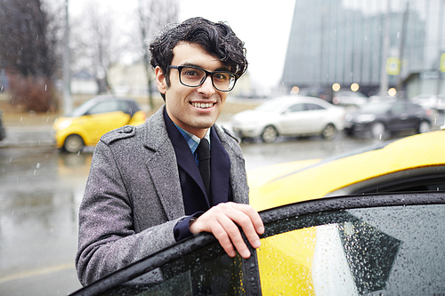 Portrait of young handsome  Middle-Eastern businessman getting in  taxi cab on rainy autumn street, smiling and  while opening car door