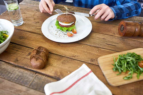Hungry human with fork and knife eating hamburger by wooden table