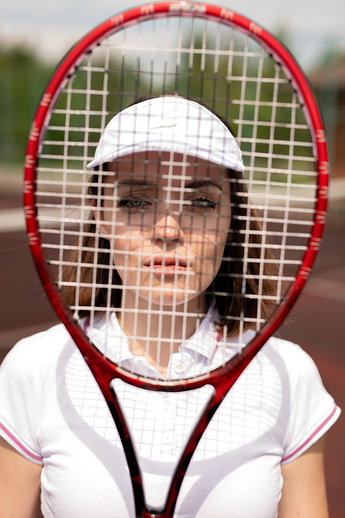Pretty young serious tennis player looking at you through racket on sunny day