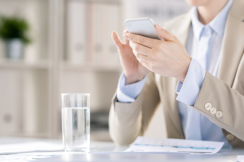 Close-up of unrecognizable businesswoman in jacket sitting at table with papers and glass of water and viewing message on smartphone