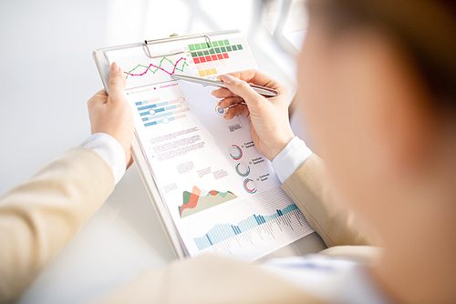 Close-up of unrecognizable busy woman pointing with pen at line chart while examining information in report