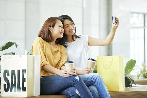 Pretty smiling young Asian women sitting on bench and taking selfie after shoppinh