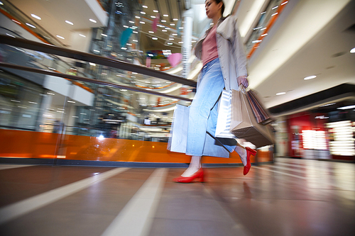 Lifestyle of shopaholic: serious young woman in coat and stylish shoes being in hurry while buying clothing on sale in mall