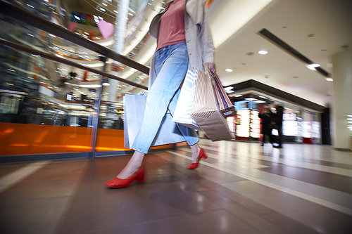 Close-up of unrecognizable young woman in spring coat and bright shoes walking over shopping mall while hurrying to buy clothing on sale