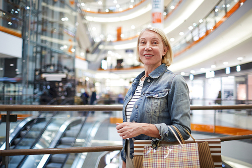 Portrait of positive beautiful mature lady with blond hair wearing denim jackets standing in spacious modern shopping mall